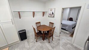 A compact room with a wooden dining table and chairs, a small trash bin, and a bed in the adjacent room. - New build 1 bed apartment with full size bathroom and kitchen. (Denham Springs)