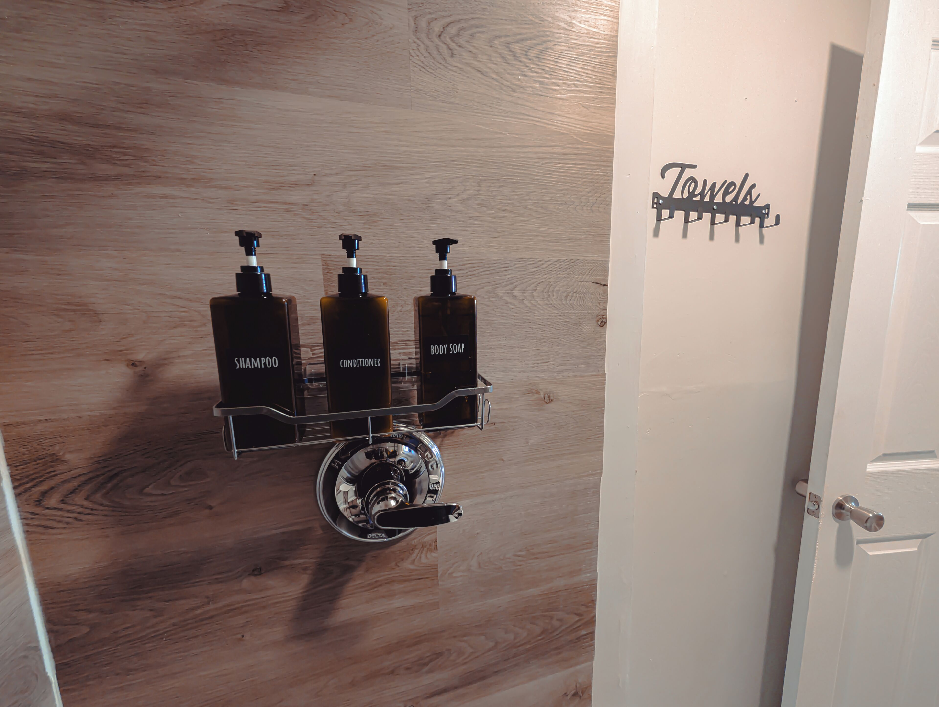 A wooden wall with a metal dispenser rack holding three bottles labeled shampoo, conditioner, and body soap.