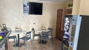 Una cocina compacta con un televisor de pantalla plana montado en la pared, un refrigerador con pantalla digital y un comedor con dos sillas y una mesa pequeña.