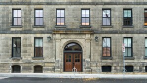 A multi-story stone building with a central arched entrance, multiple windows, and a street in front.