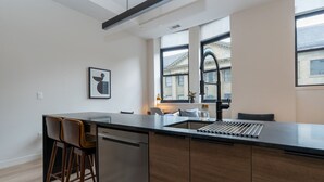 A modern kitchen with a large island, bar stools, a built-in dishwasher, and a window with a view of buildings.