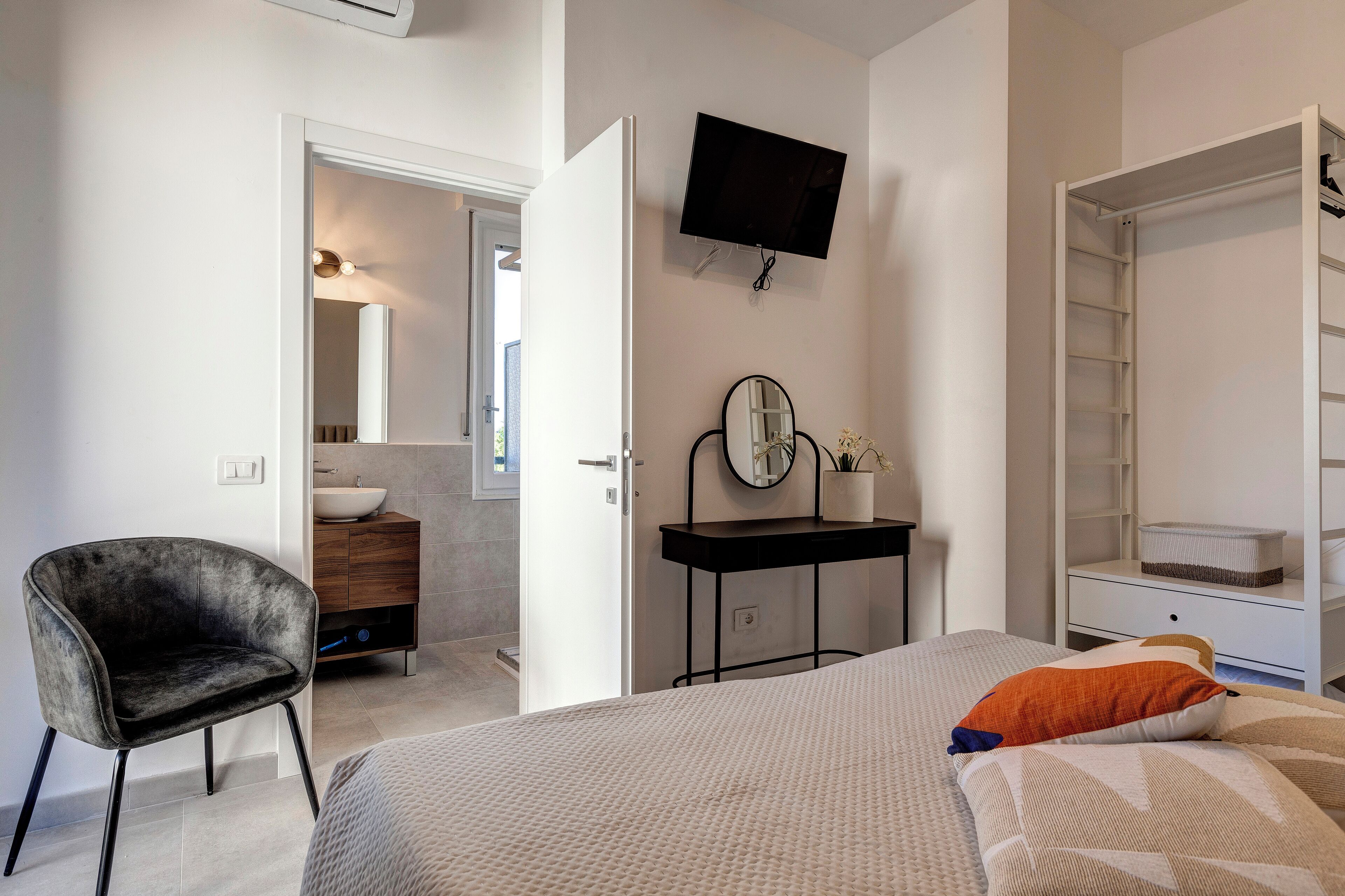 Un dormitorio con cama, una silla, tocador con espejo, televisor y armario.