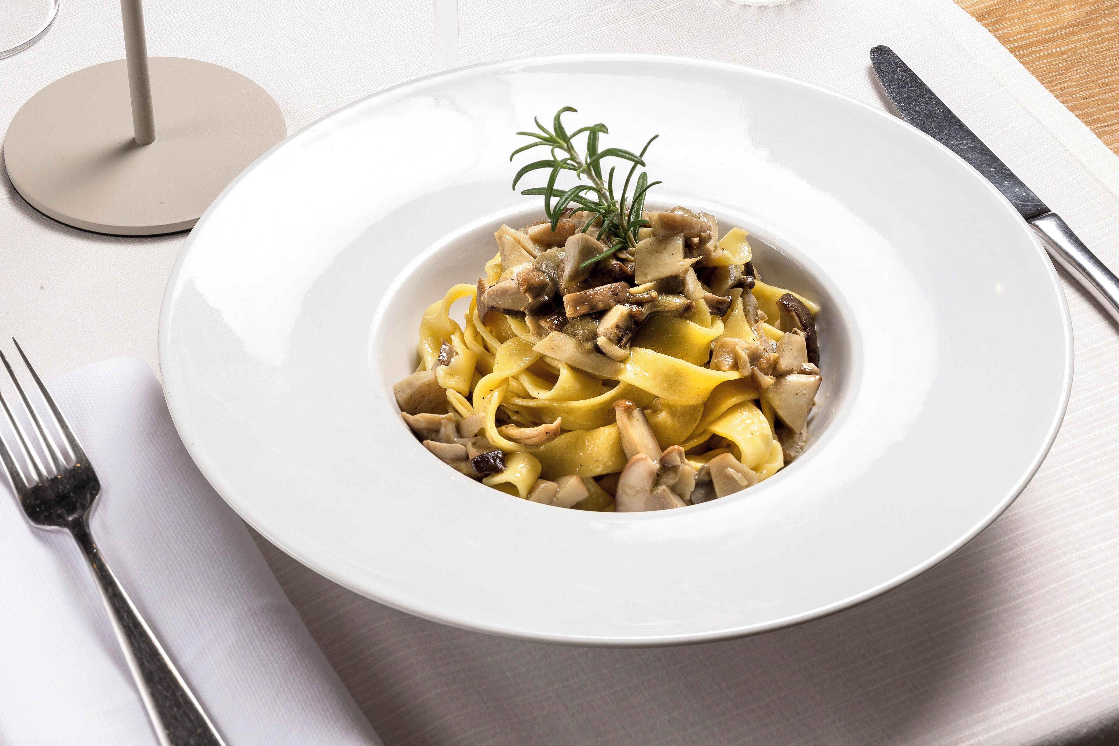 A white plate of pasta with mushrooms and a sprig of rosemary on a white tablecloth.