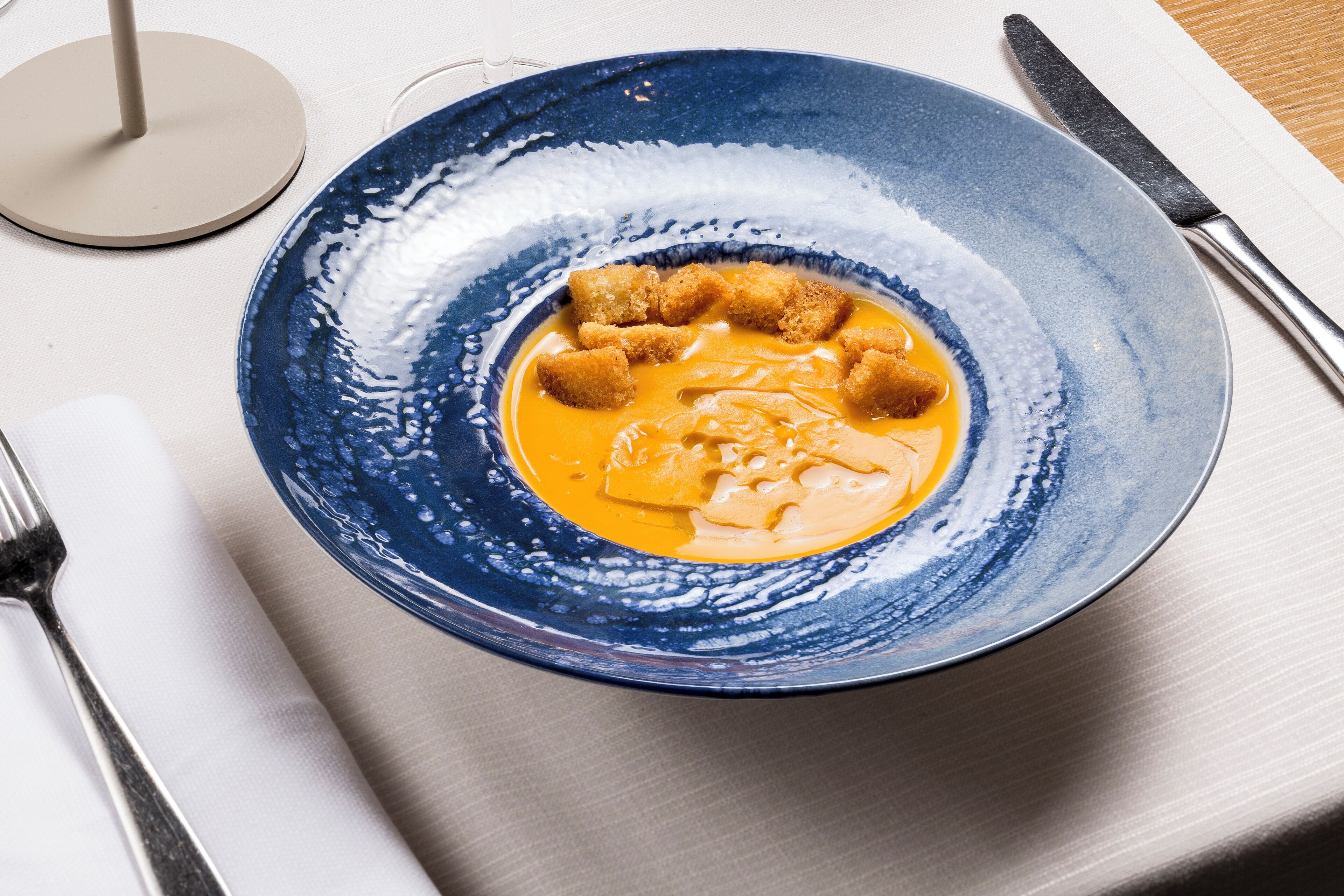 Eine blaue Keramikschale mit cremig-orangem Dip und Croutons auf einer weißen Tischdecke.