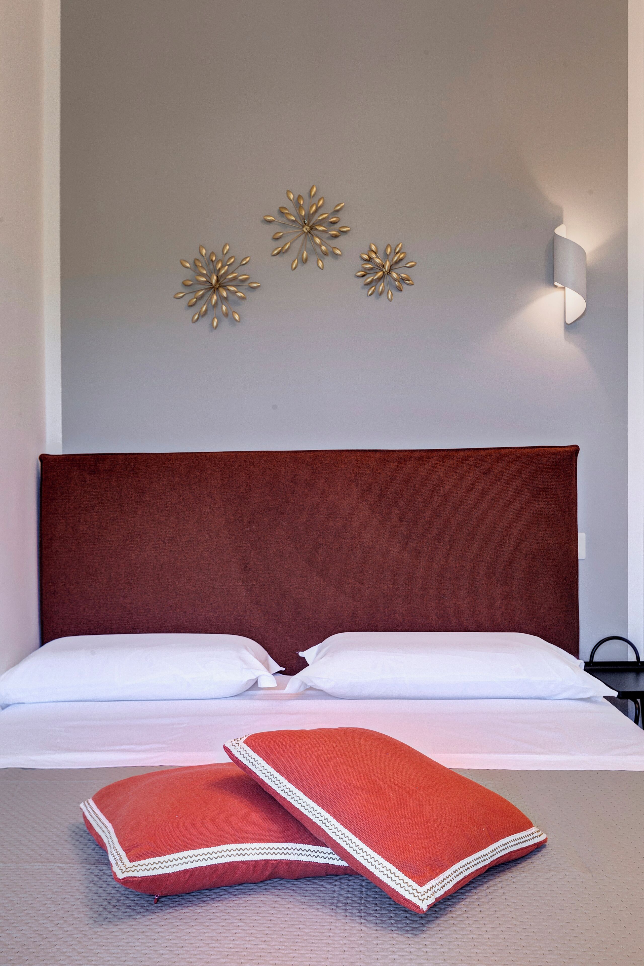 Een net opgemaakt bed met een rode hoofdbord, witte kussens en twee rode kussens met witte bies. Daarboven hangen drie decoratieve items aan de muur.