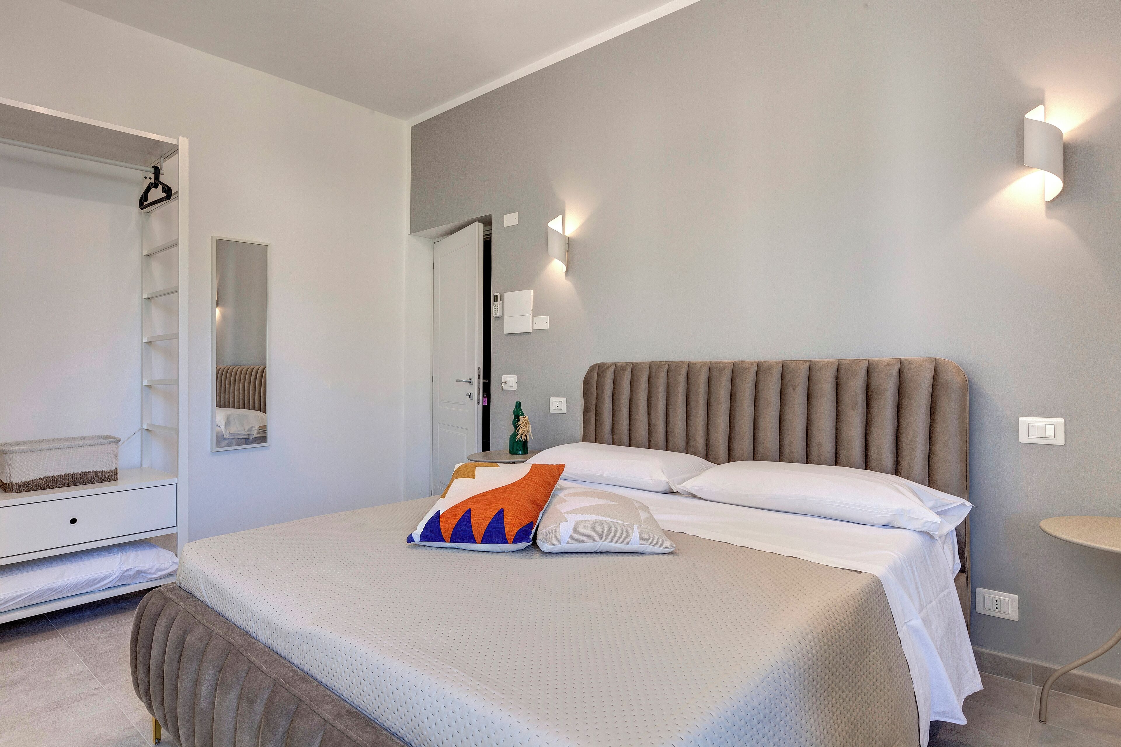 Une chambre à coucher comprenant un lit, une table de chevet, un miroir, une armoire et un luminaire fixé au mur.