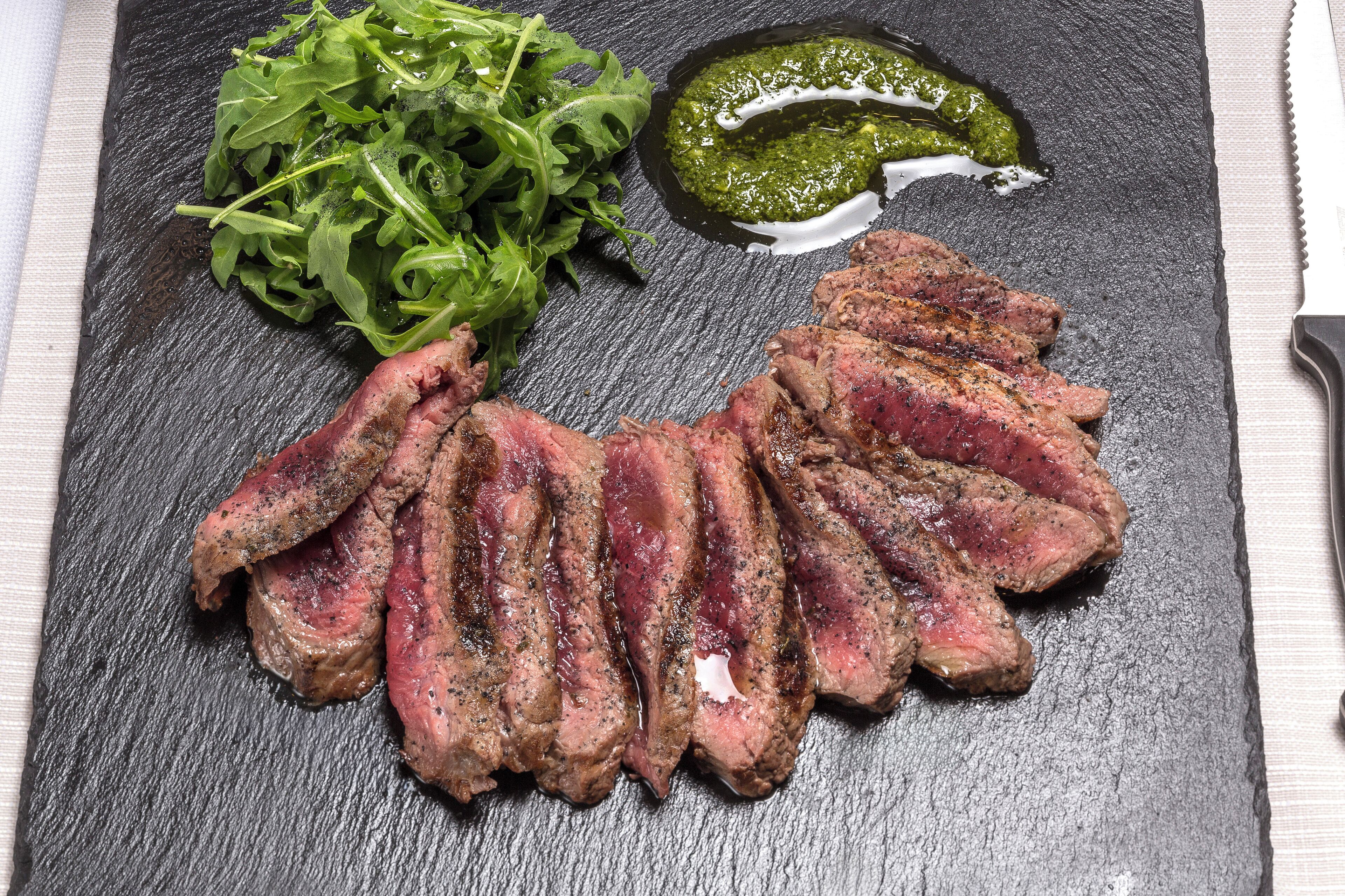 A steak with greens and a sauce on a slate board.