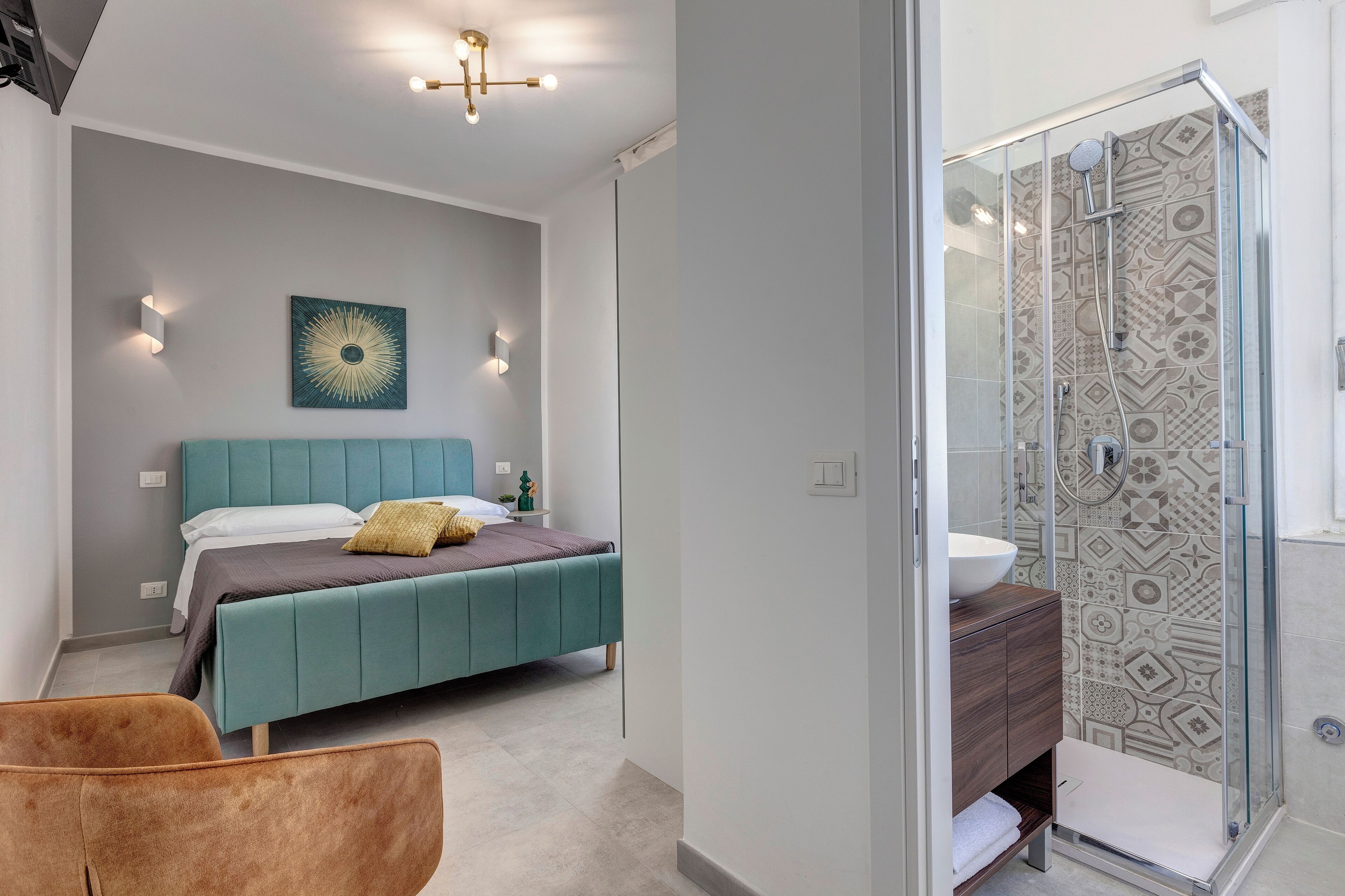 Une chambre moderne avec un lit de couleur turquoise, un fauteuil marron et une salle de bain aux carreaux décoratifs et dotée d’une douche en verre.