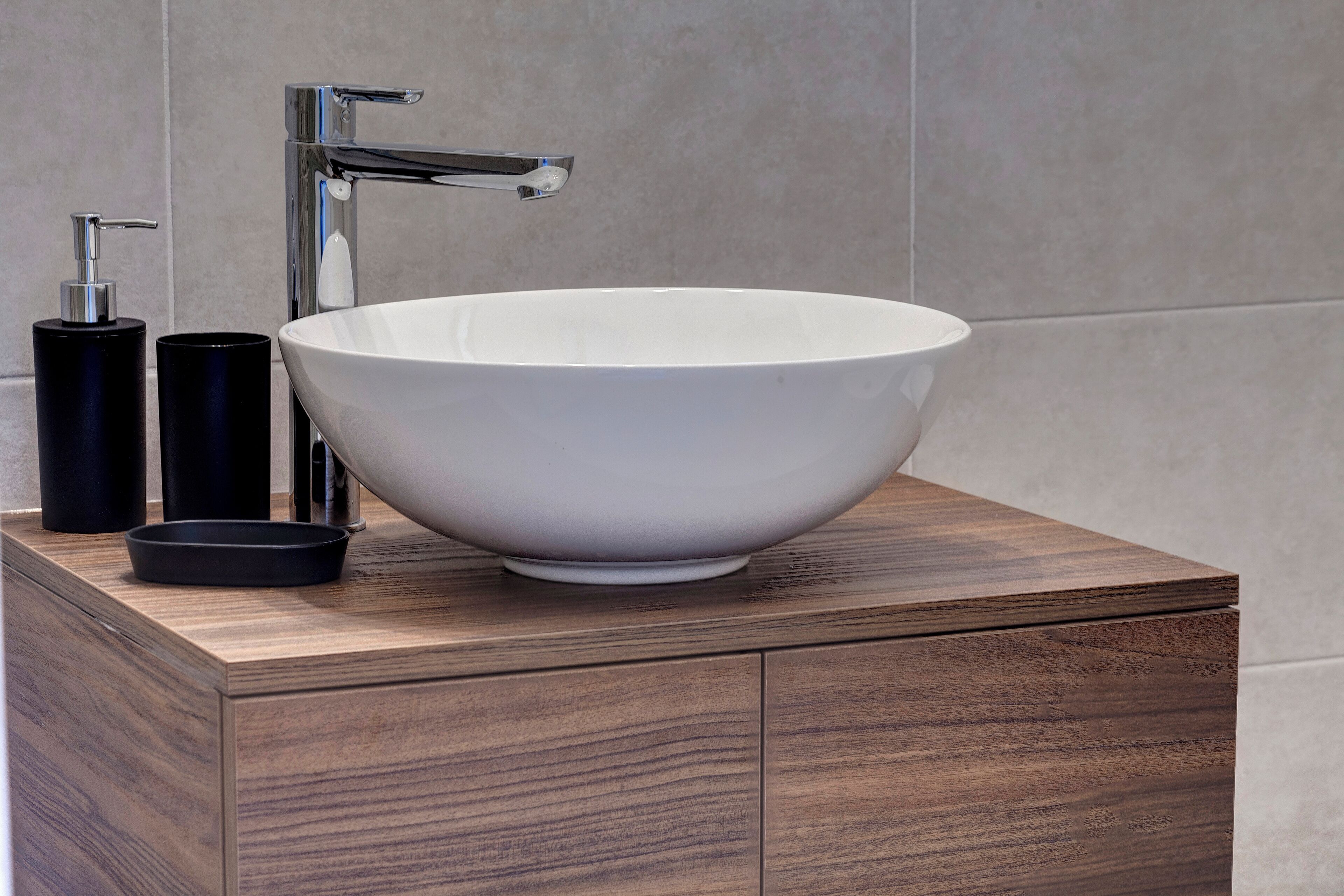 A modern bathroom with a large white vessel sink, a chrome faucet, and black toiletries on a wooden vanity.