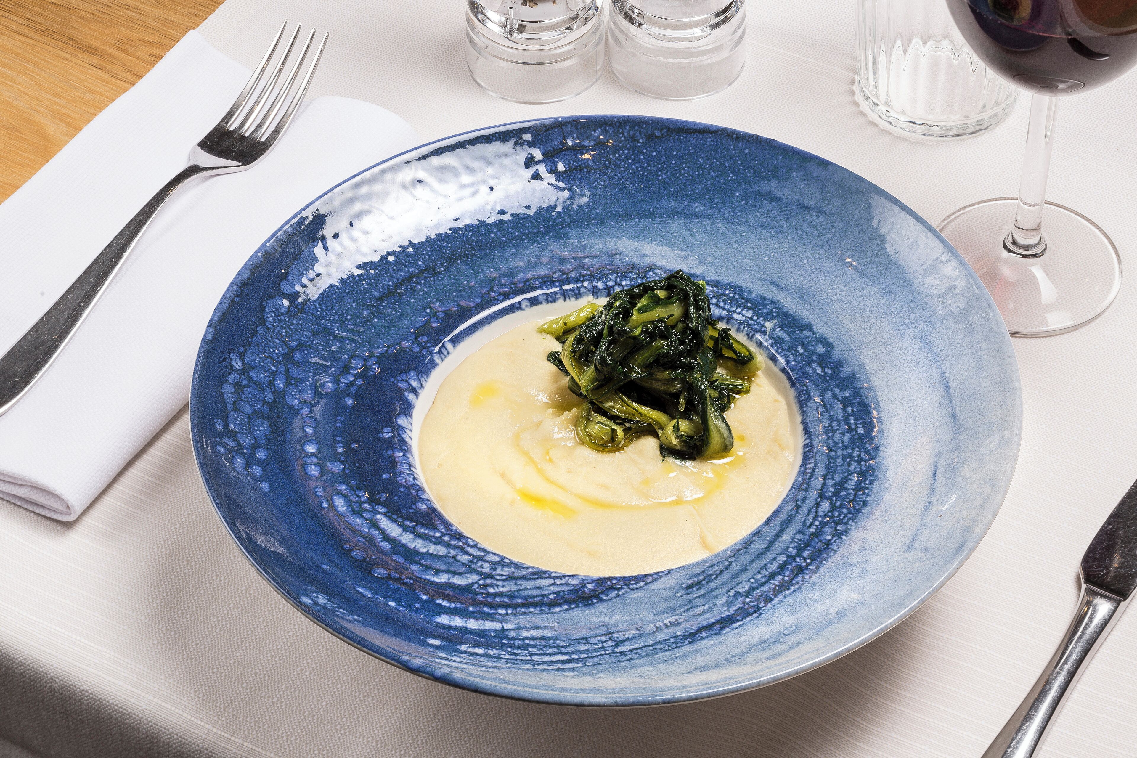 Une assiette en céramique bleue, garnie d’une sauce crémeuse et de légumes verts, accompagnée d’une fourchette et d’un verre de vin rouge, le tout posé sur une table.