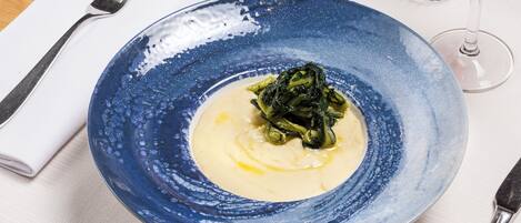 Une assiette en céramique bleue, garnie d’une sauce crémeuse et de légumes verts, accompagnée d’une fourchette et d’un verre de vin rouge, le tout posé sur une table.