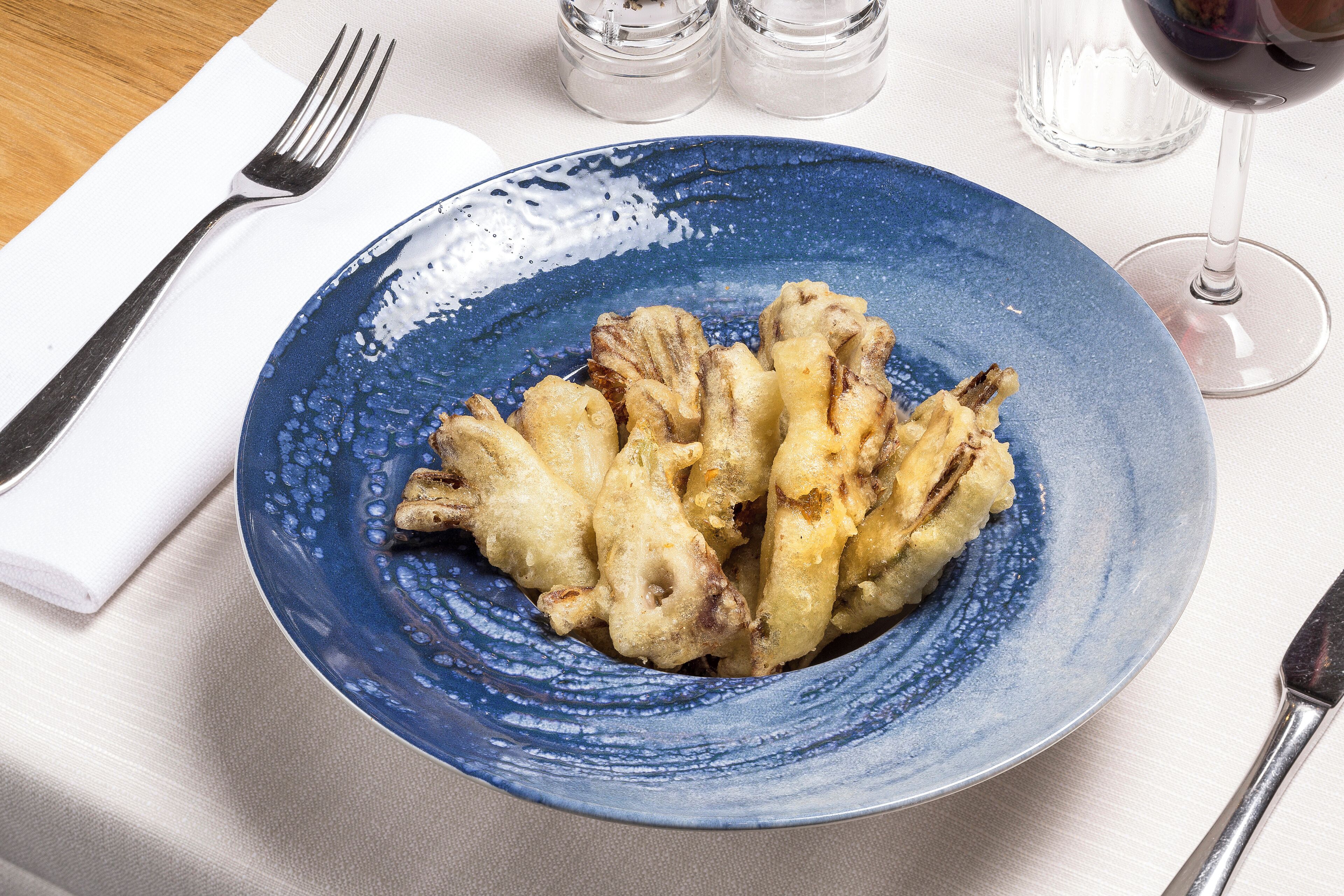 A blue plate with fried food on a table with a fork and a wine glass.