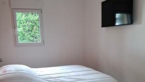 A bedroom with a bed, a window, a TV mounted on the wall, and a small table with a chair.