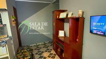 Uma área de recepção com piso ladrilhado, uma estante de madeira, uma televisão e uma placa fixada na parede.
