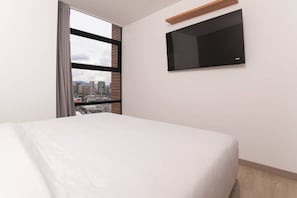 A hotel room with a bed, a TV mounted on the wall, and a window with a city view.