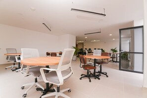 A modern conference room with round tables and white mesh chairs, equipped with overhead lighting and potted plants.