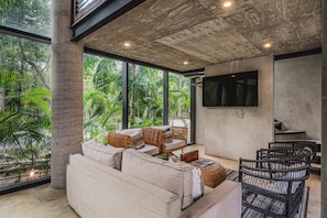 A modern living room with a sofa, wicker furniture, a flat-screen TV, and large windows overlooking greenery. - 2BR Penthouse w Private Rooftop | Jacuzzi & Gym Access (Tulum)