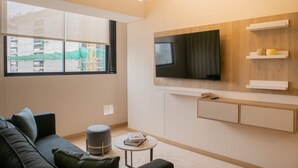 A modern living room with a large TV, built-in shelves, a sofa, a small round table, and a window with a view of a construction site. - w Cozy Bright 2BR Stay in Barranco (Lima)