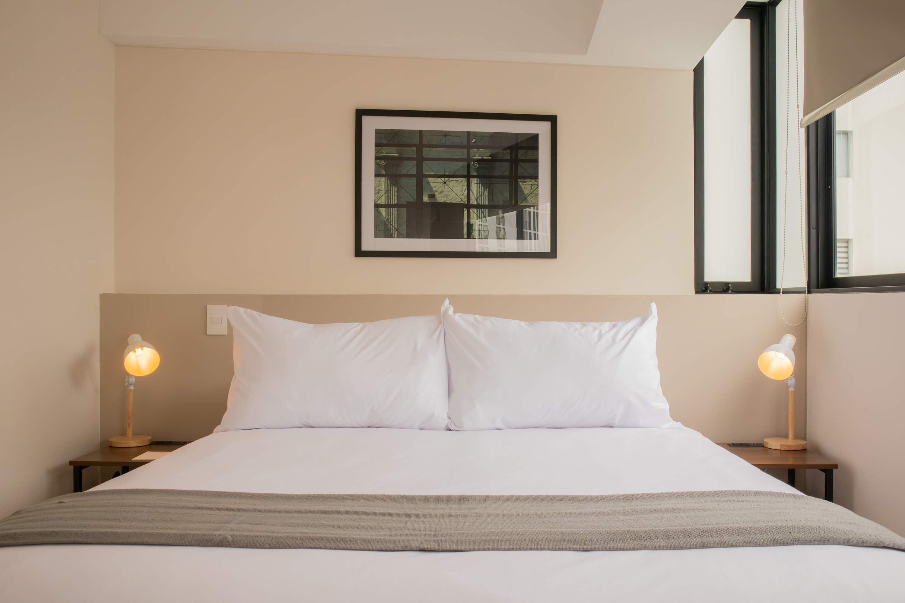 A neatly made bed with white linens, two bedside lamps, and a framed window above.