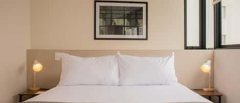 A neatly made bed with white linens, two bedside lamps, and a framed window above.