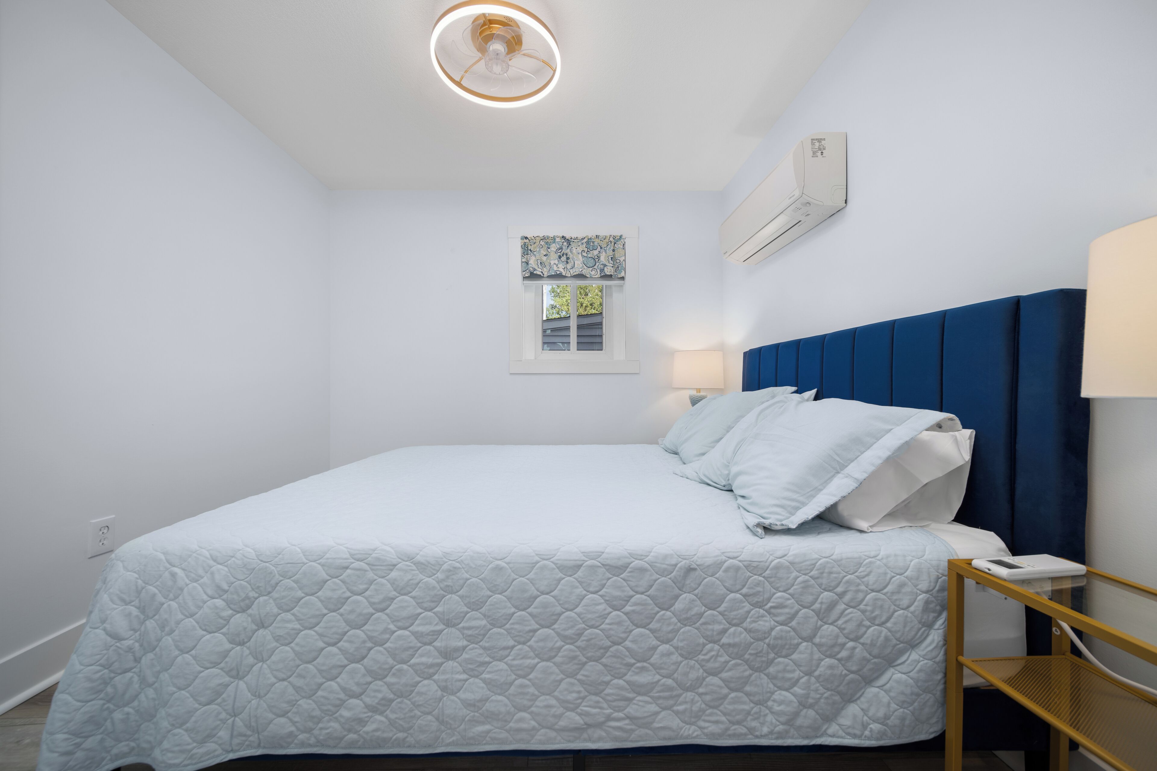 Ein ordentlich bezogenes Bett mit weißer gesteppter Tagesdecke, einem blauen Kopfteil und einem Nachttisch mit Lampe.