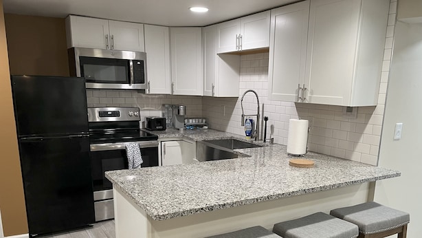 A kitchen with white cabinets, stainless steel appliances, a granite countertop, and three bar stools. - Mid-Century Modern Broad Ripple Stay with Style, Comfort & Local Color (Indianapolis)