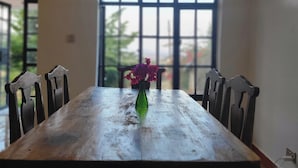 A dining table with chairs, a vase with flowers, and a window with a view of greenery. - PUMZIKA HILL CAMPSITE (Kisongo)