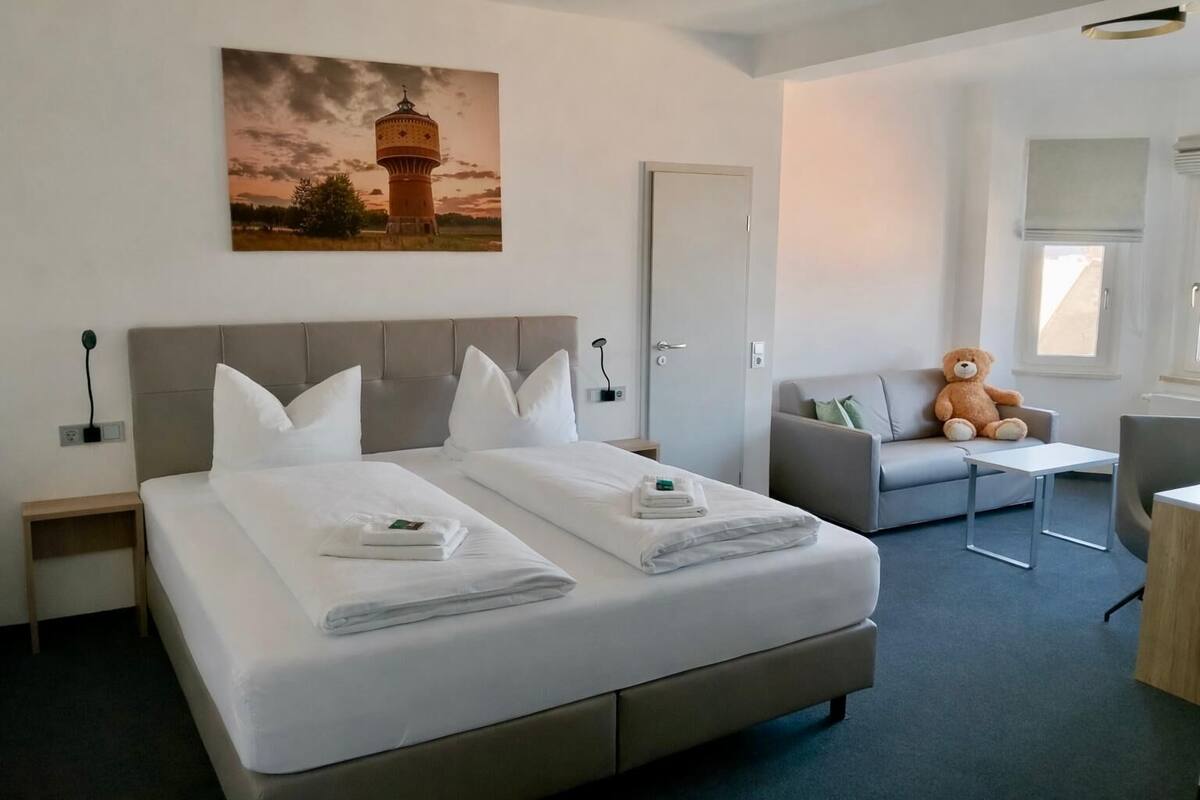 A hotel room with a large bed, a sofa, a chair, a teddy bear, and a painting on the wall.