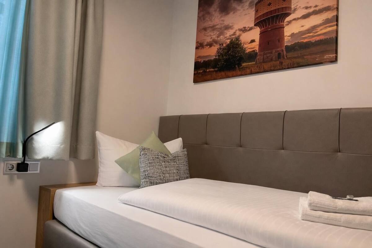 A neatly made bed with a headboard, a bedside lamp, and a painting of a tower on the wall.