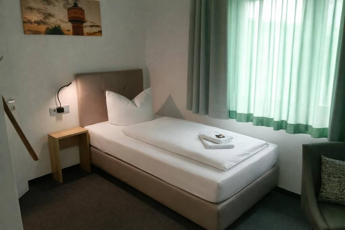 A single bed with a headboard, a bedside table, a wall-mounted light, a window with curtains, and a painting of a lighthouse.