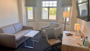 A hotel room with a sofa, two armchairs, a small table, a desk with a lamp, and a television.