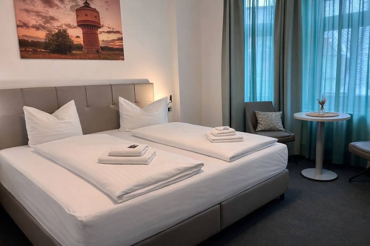 A hotel room with a large bed, two pillows, a small round table with a vase, and a chair.
