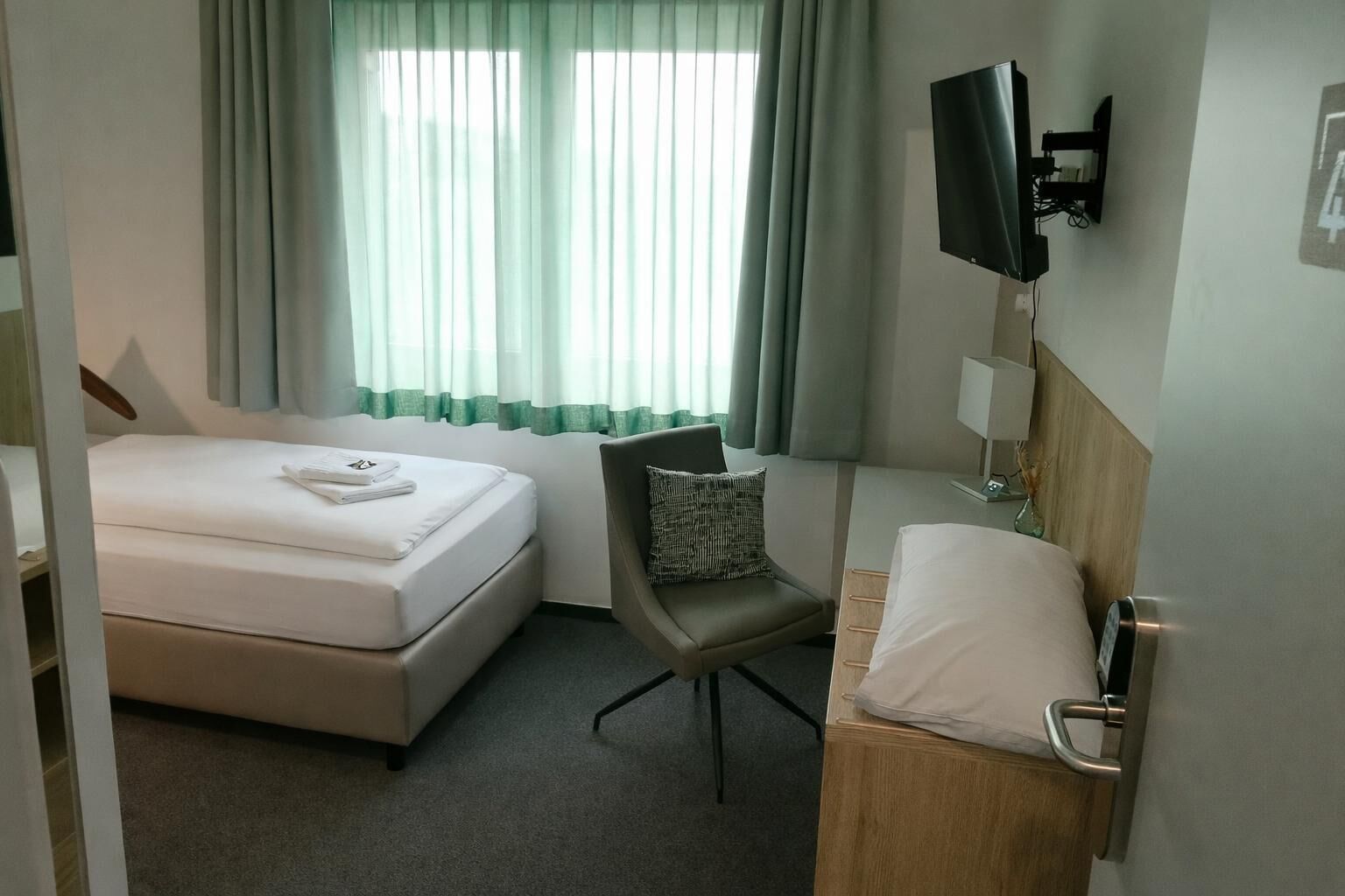 A hotel room with a bed, a chair, a TV mounted on the wall, and a bedside table with a lamp.