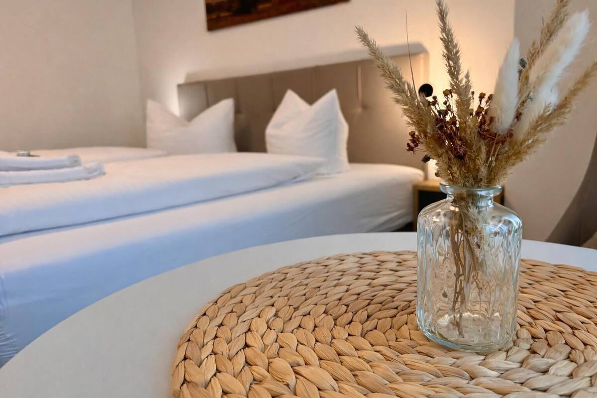 A hotel room with a bed, a bedside table with a vase of dried plants, and a woven placemat.
