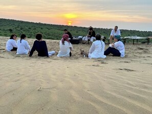 A group of people sitting on a sandy beach at sunset. - Royal Humsafar Hostel Jaisalmer (Jaisalmer)