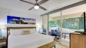 A modern hotel room with a large bed, a ceiling fan, and a view of a garden through a sliding glass door.