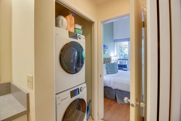 Un cuarto de lavado con lavadoras y secadoras apiladas, una encimera y una puerta que da a un dormitorio con una cama y una ventana.