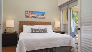 A neatly made bed with a pillow labeled "BEACH," a wall-mounted painting of a beach scene, and a window with a view of the outdoors.