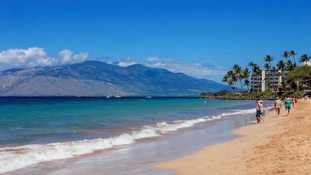 A beach with clear blue water, a sandy shore, and a mountain in the background.