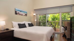 Un dormitorio con una cama grande, una mesita de noche, una lámpara, un cuadro de una playa y vistas a palmeras a través de la ventana.