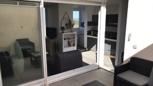 A sunlit patio with a sliding glass door leading to a living area, furnished with a sofa, a coffee table, and a TV. - Studio on the port of Carnon with patio, 5mn from the beach, ideal for 3 people (Mauguio)