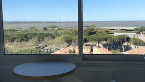 A room with a large window offering a view of a river and greenery, a round table, and two white chairs. - Studio on the port of Carnon with patio, 5mn from the beach, ideal for 3 people (Mauguio)