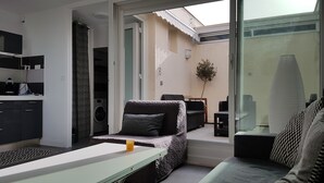 A modern living room with a sofa, a small table, and a kitchen area with a washing machine. - Studio on the port of Carnon with patio, 5mn from the beach, ideal for 3 people (Mauguio)