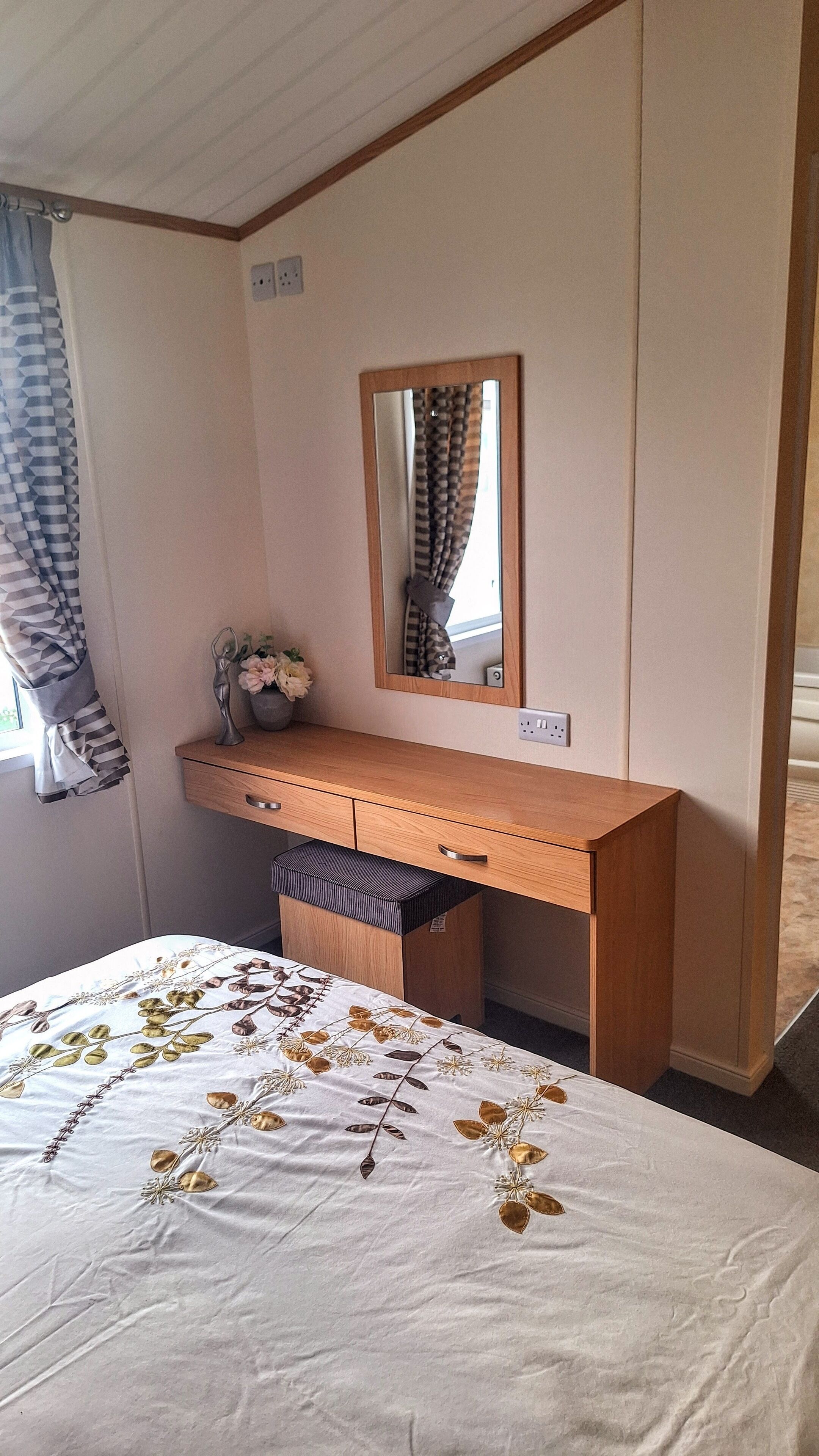 A bedroom with a wooden desk, a mirror, a bed with a floral bedspread, and a window with curtains.