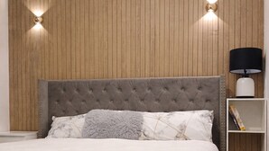 Een slaapkamer met een geborduurde hoofdbord, een witte dekbedovertrek, een grijs zacht kussen, een wit kussen, een nachtkastje met een lamp en een houten wandpaneel.