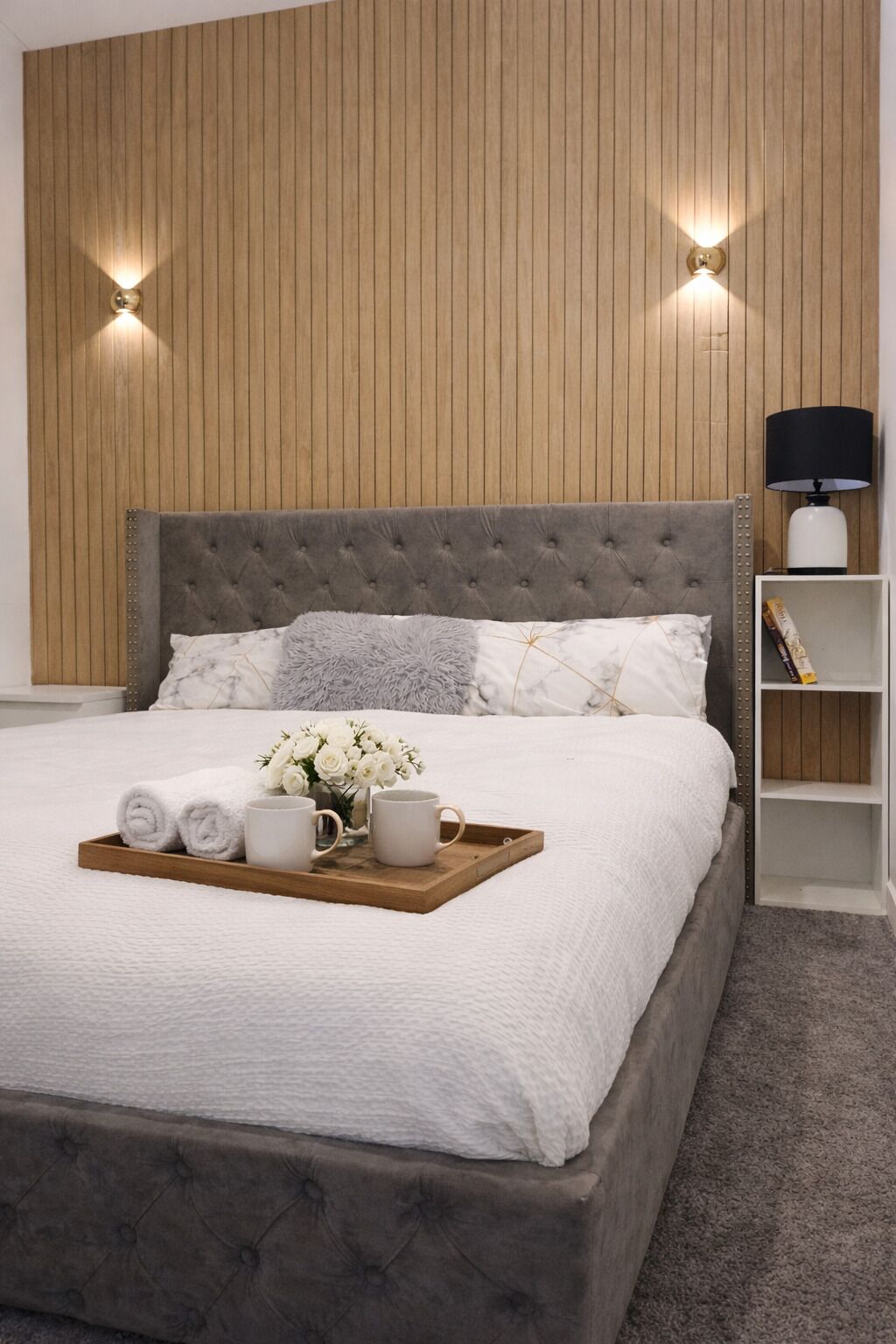 A bedroom with a tufted headboard, a wooden panel wall, two wall-mounted lights, a bedside table with a lamp, and a tray with towels, a vase of flowers, and two mugs.