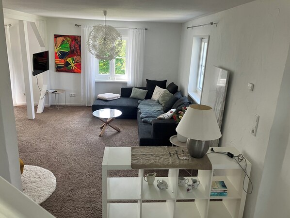 A modern living room with a sofa, a coffee table, and a TV. There is a window with curtains, a painting on the wall, and a lamp on a side table. - Beautiful country house in the Rhön with a large garden (Zeitlofs)