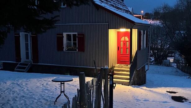 Et træhus med rødt dør, sneklædt tag og en lille veranda.