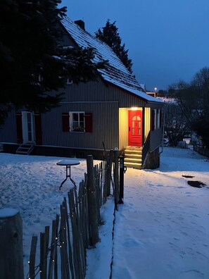 A wooden house with a red door, a snow-covered roof, and a small porch area. - Beautiful country house in the Rhön with a large garden (Zeitlofs)
