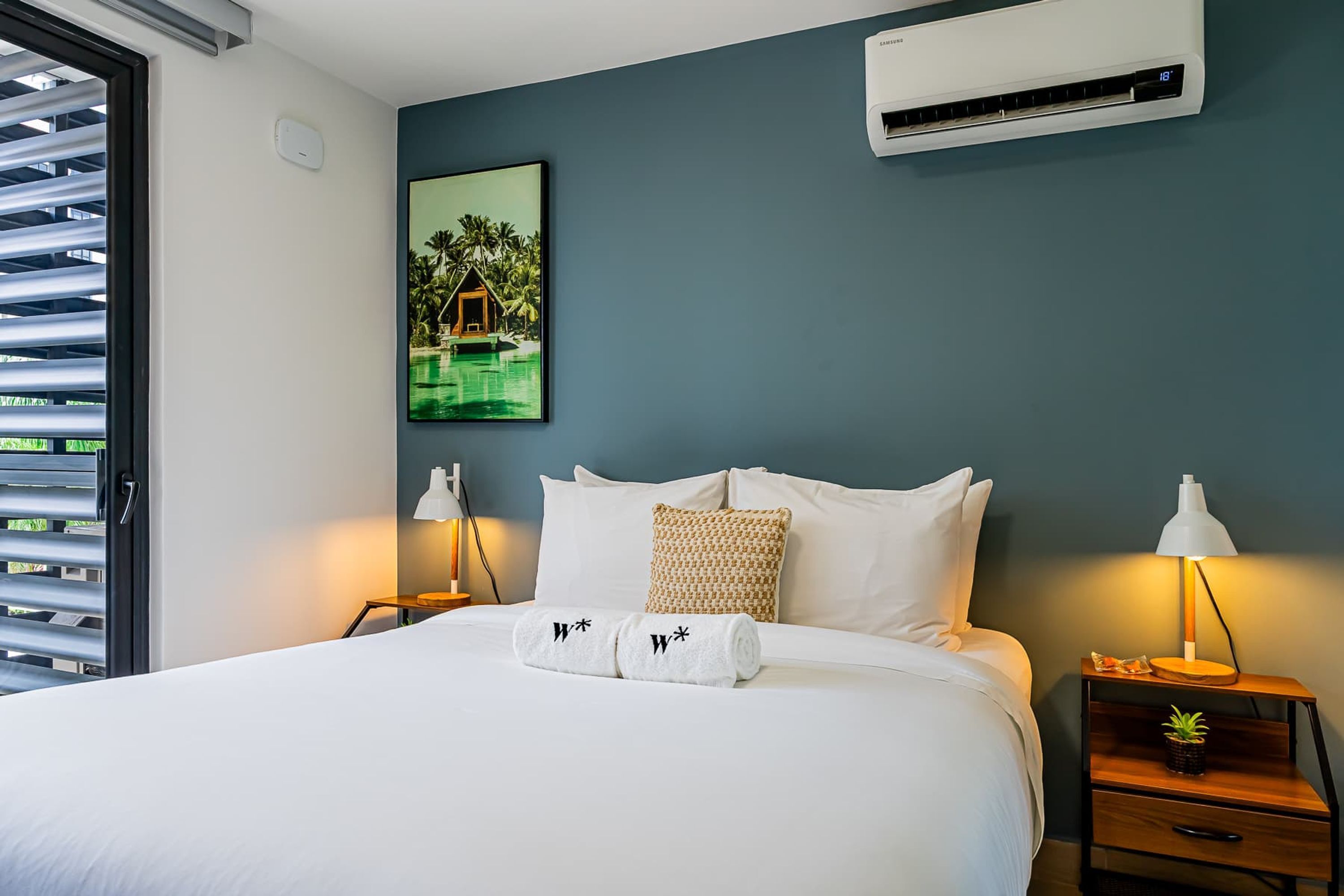 A neatly made bed with white linens and two towels with a logo, a wall-mounted air conditioner, a framed picture of a tropical resort, and a bedside table with a lamp and a small plant.